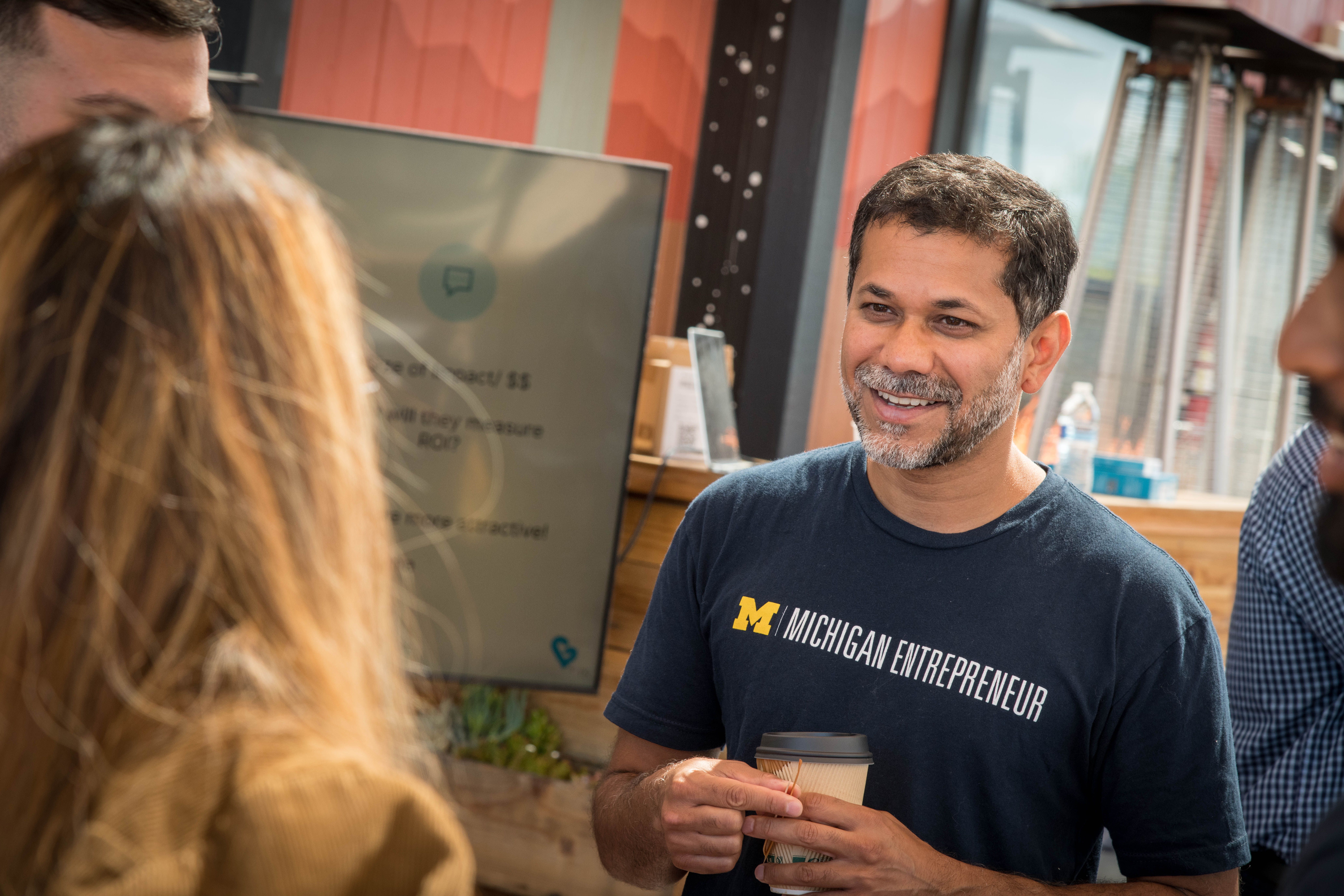 Inder Singh smiles while holding a cup of coffee and speaking with a trek student