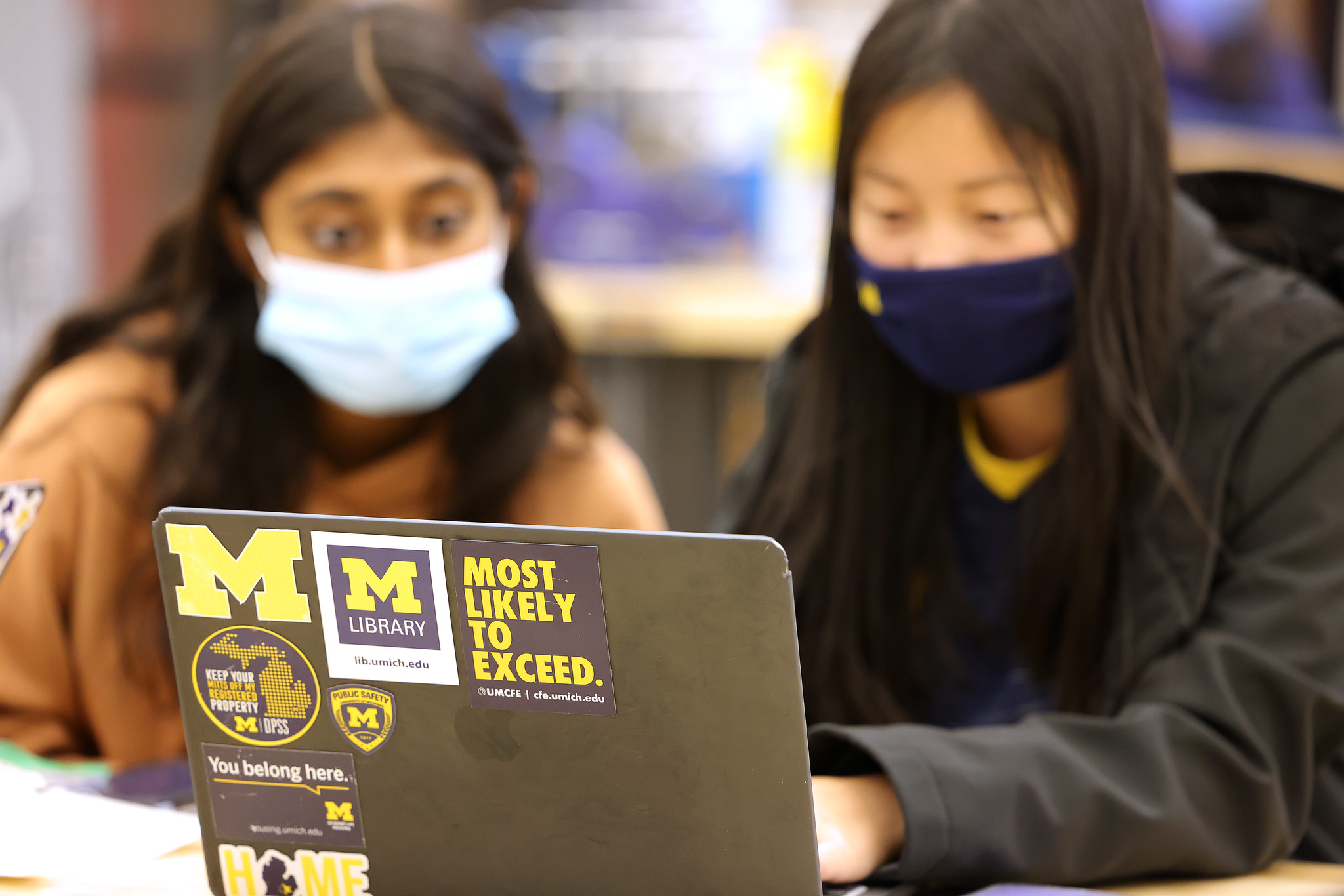 Two female University of Michigan students in masks collaborate around a laptop with a Most LIkely To Exceed Sticker