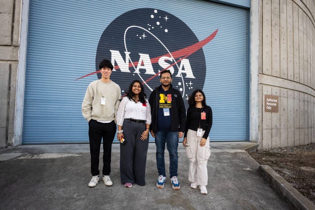 A group of four students pose in front of a mural of the NASA logo.