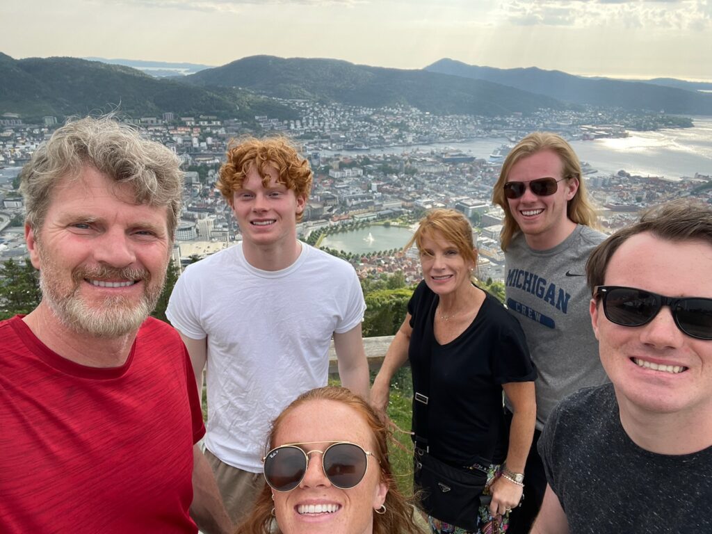 Chris poses with his family on a trip to Norway. They are smiling with a spectacular view of maountains and city scenery behind them.