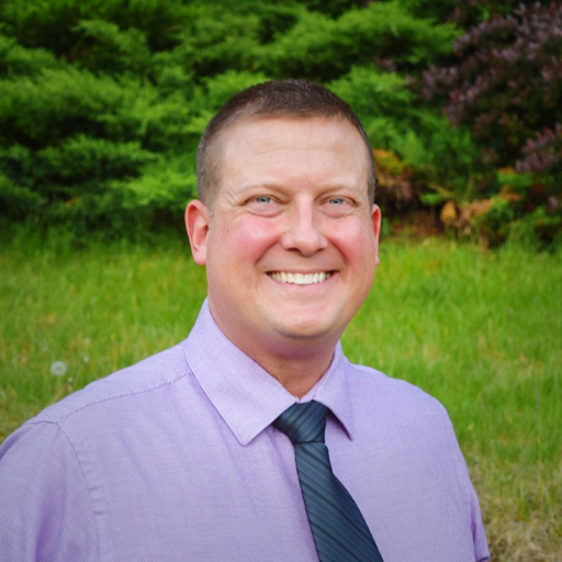 david brewer headshot, smiling wearing a purple shirt with a blue tie and a scenic background with green grass & trees