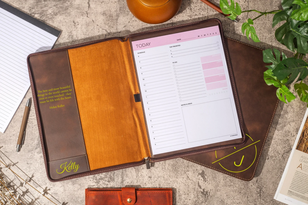 A leather planner sits open on a table. There is an inspirational quote and personalized name on the left hand side. On the right side, a daily planner page shows daily schedule, top priorities, a to-do list, and notes and ideas. 