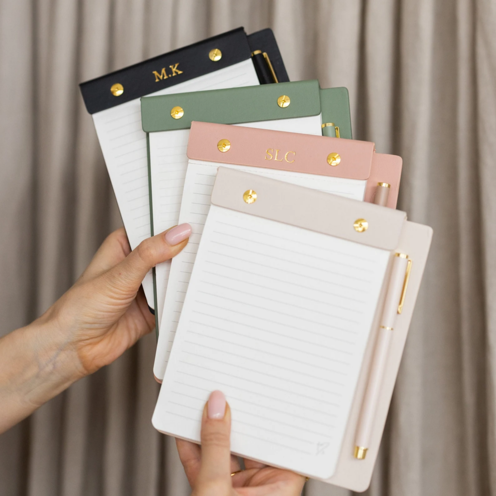 Custom notepad is shown in four colors, black, green, pink, and cream. Each notepad holder contains a spot for a matching pen.