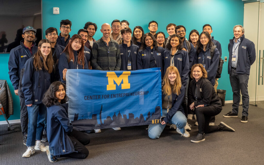 CFE trek students pose with a flag that displays a block M and the text "Center for Entrepreneurship. NEXT."