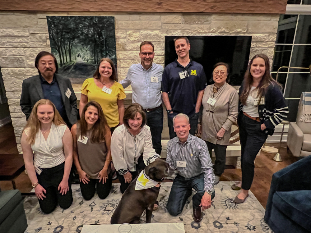group of U-mich alumni and students around a brick fireplace wall smiling at the camera