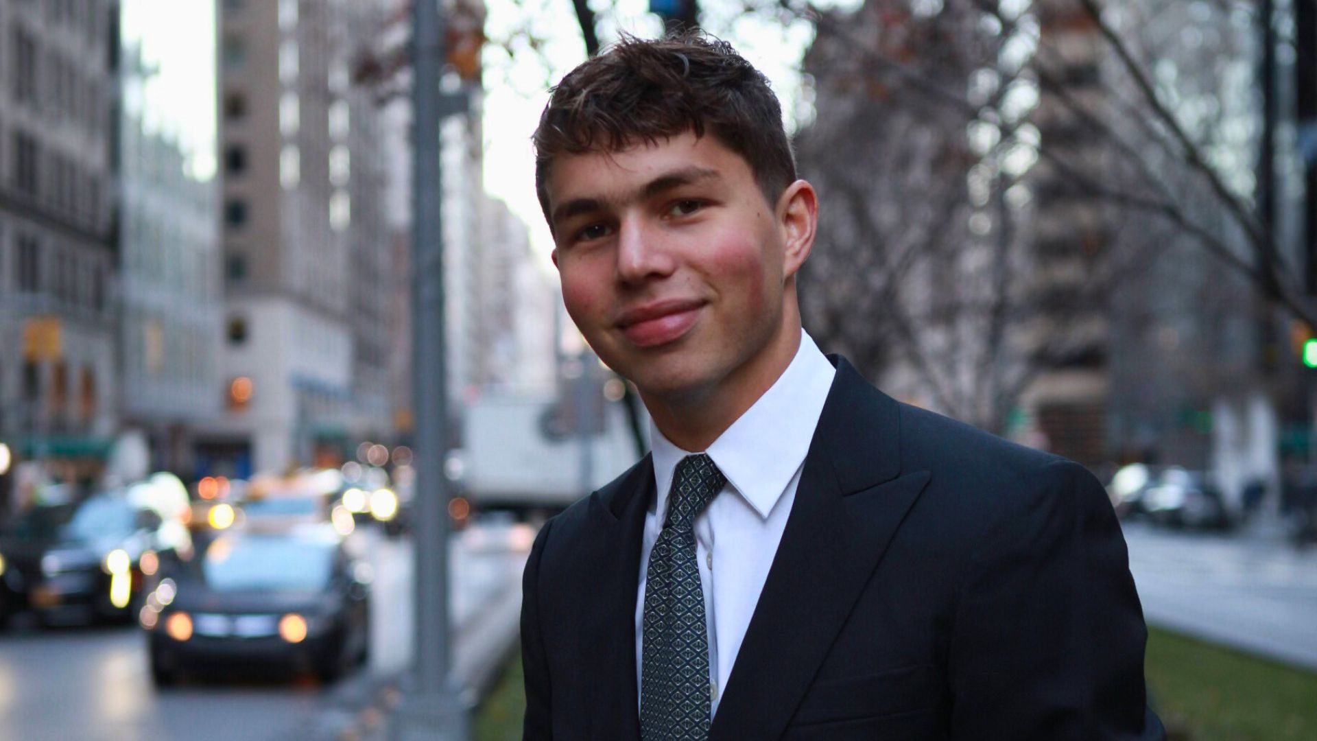 David poses in a black suit and tie against a bustling city backdrop.