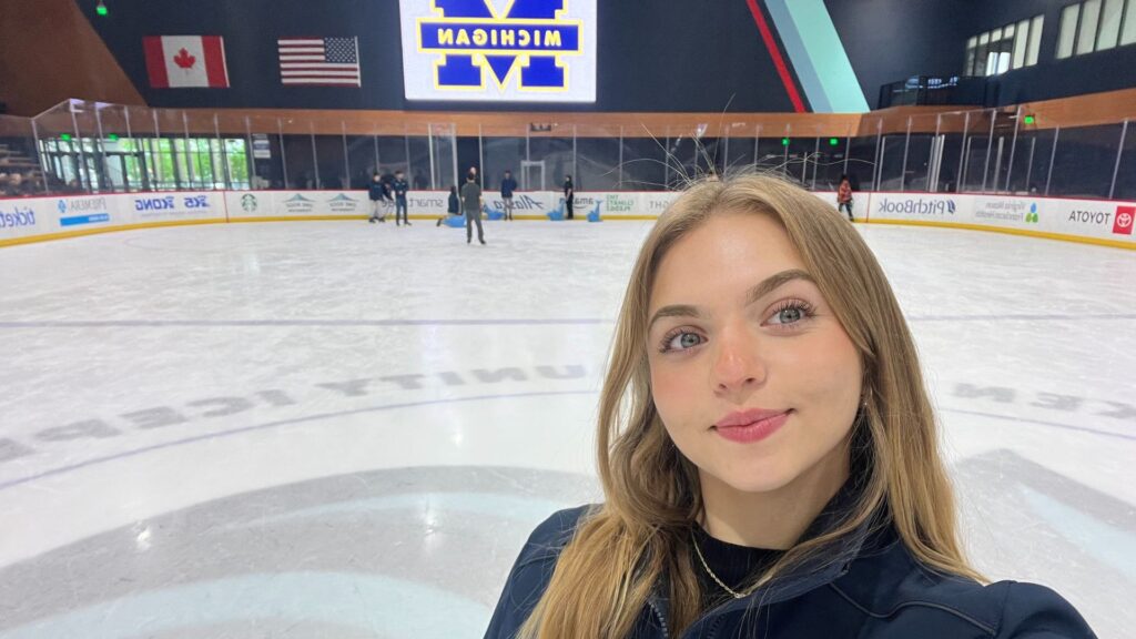 Isabella poses at an ice rink in front of a big screen featuring the Block M.