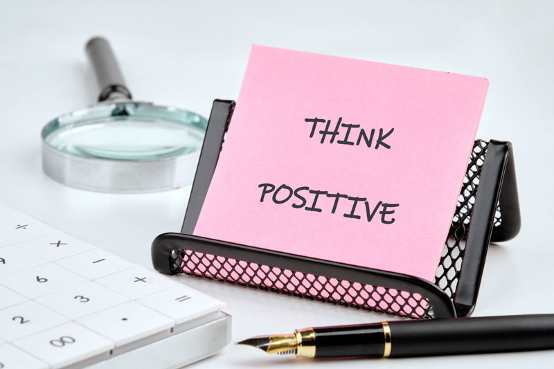 A pink sticky note propped in a black holder says THINK POSITIVE. Nearby, a calculator, pen, and magnifier suggest financial planning and a focus on achieving goals in an office setting.