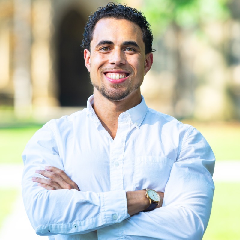 Marc Howland smiles for a headshot against an outdoor background. He is wearing a light blue button-up shirt.