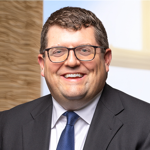 mike turner headshot. A man with a side part in his brown hair wearing glasses and a blazer with a blue tie