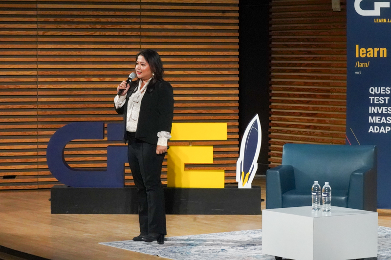 Priyi Gogoi on the EHour stage with the large CFE maize and blue signage in the background. She wears a black suit and is holding a microphone
