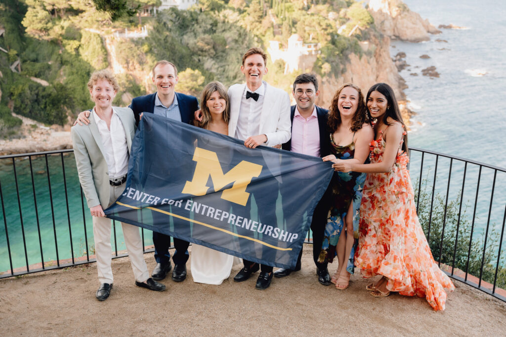 Emma and Daniil are shown at their wedding. They are posing with fellow ELP students and holding up a navy flag that reads "Center for Entrepreneurship" with a Block M.