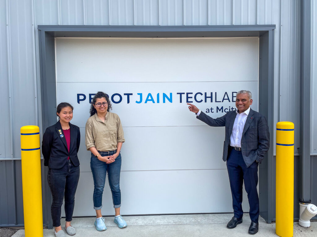 Two female students stand outside the garage doors of the Perot Jain TechLab at mcity garages with Anurag Jain of Perot Jain