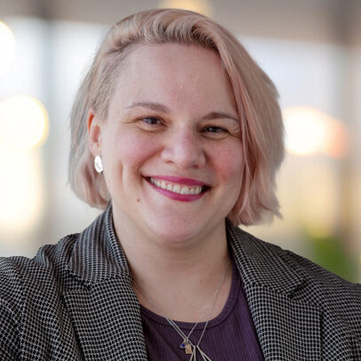 Emily demko is a woman with short blond hair. She is wearing a white and black checked blazer with a purple shirt and gold jewlery