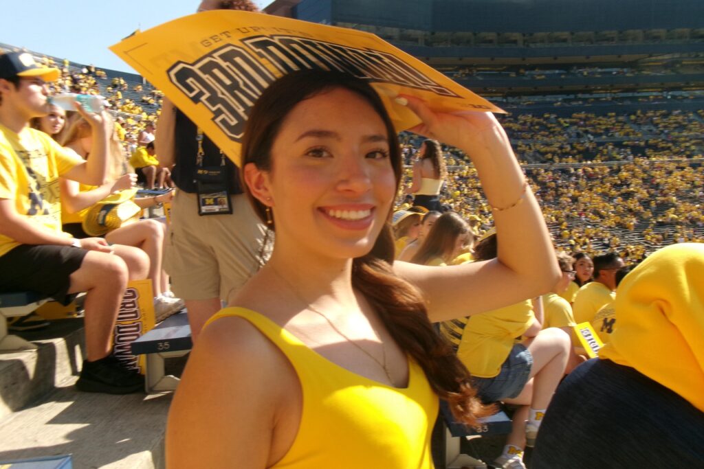 Ivana smiles while holding a sign. She is in the stands at a Michigan football game, surrounded by a sea of Maize.