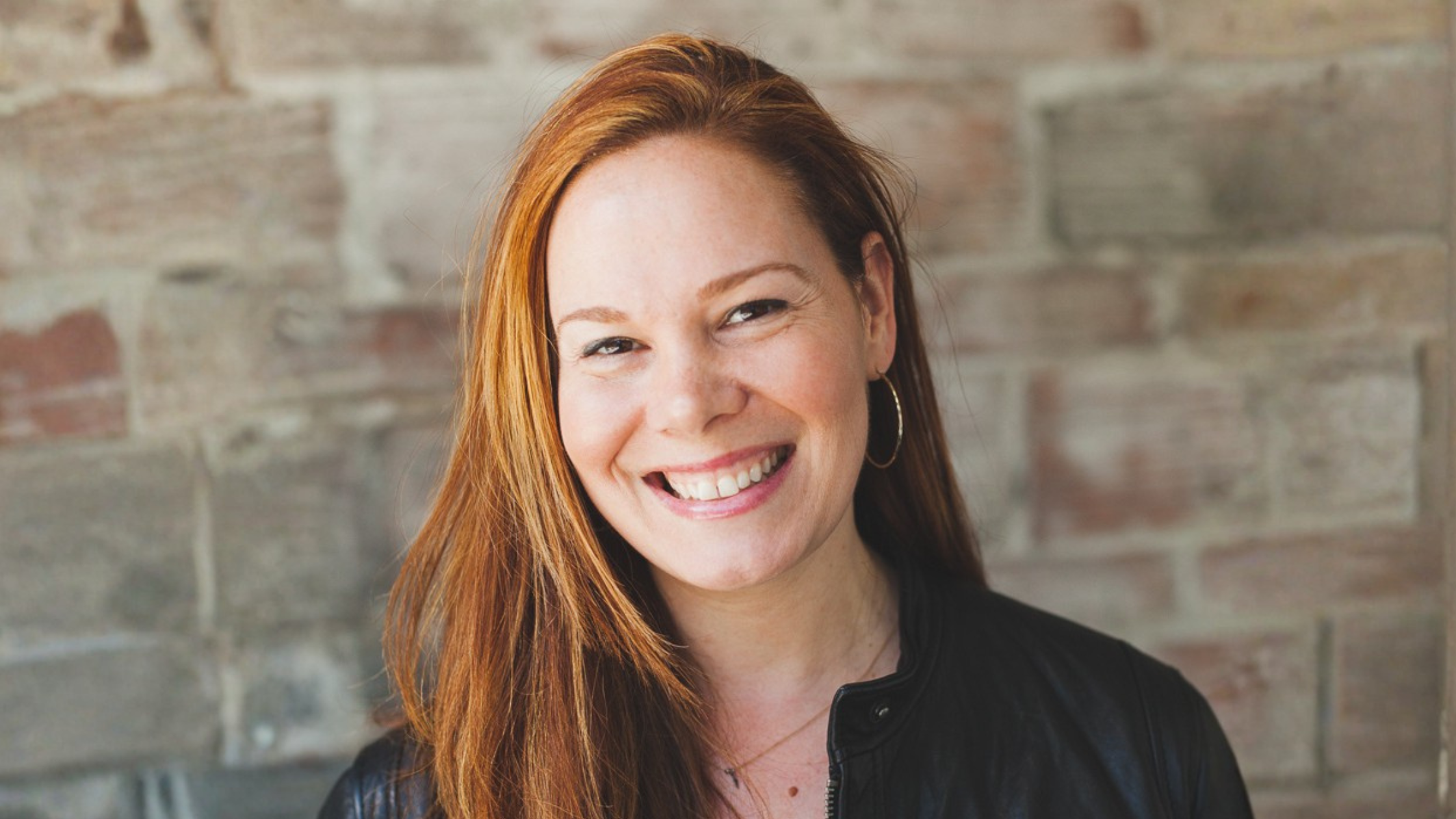 Kristin Toth smiles while posing for a headshot. She has long red hair, and she is standing in front of a brick background.