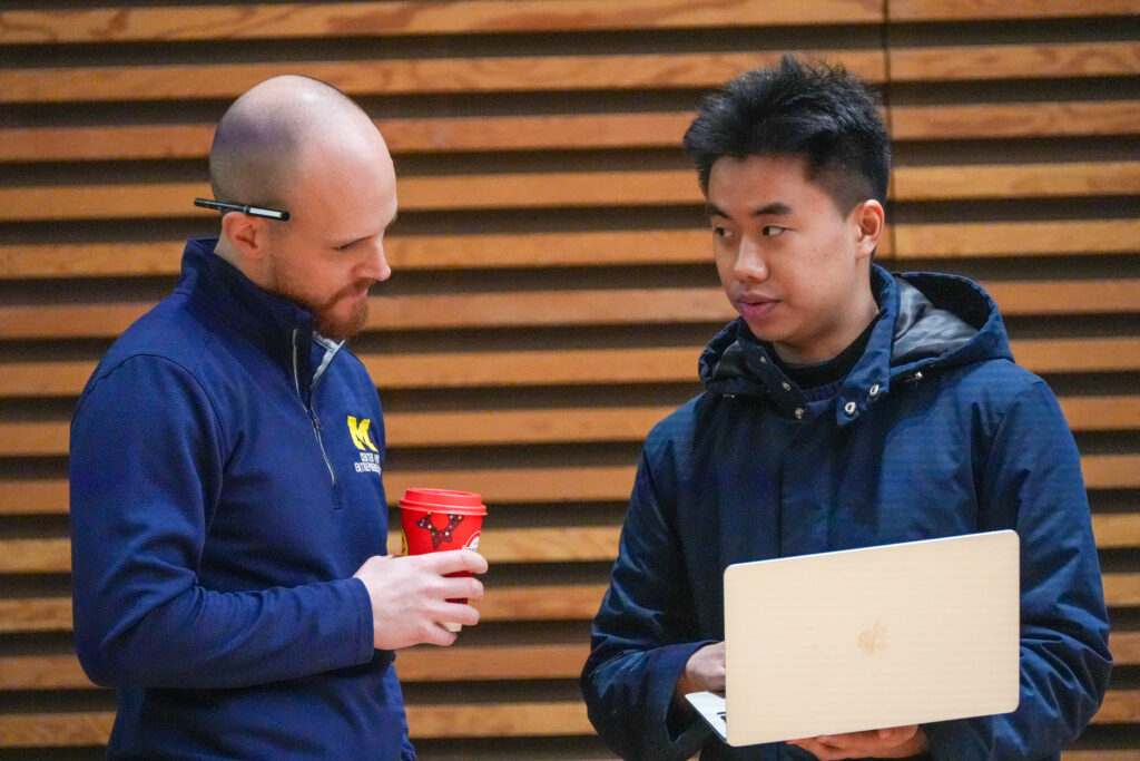 Eric speaks with a student after class. He is holding a coffee and the student is showing him something on their laptop.