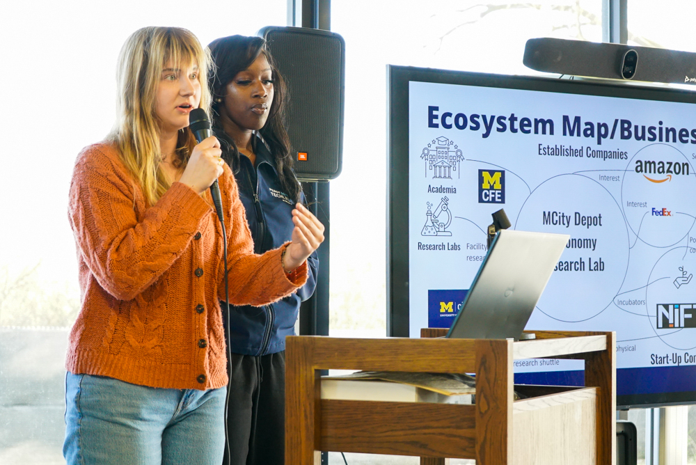 Two women one with blond hair wearing a button up orange sweater, the other with long dark hair wearing a blue Perot Jain TechLab Mcity jacket giving a presentation