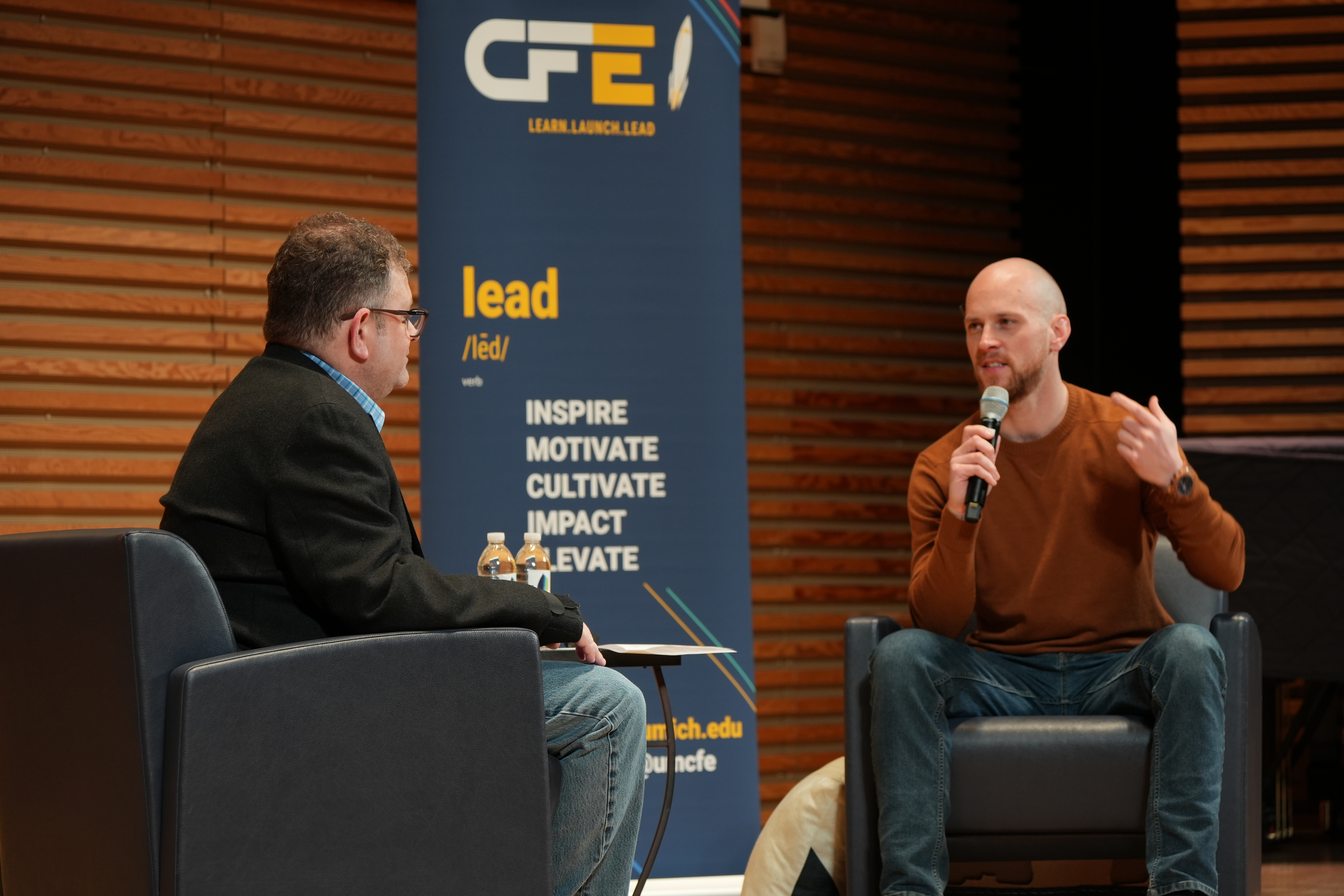 Eric sits in a chair across from an EHour speaker. He holds a microphone and speaks passionately to students as he interviews the special guest.