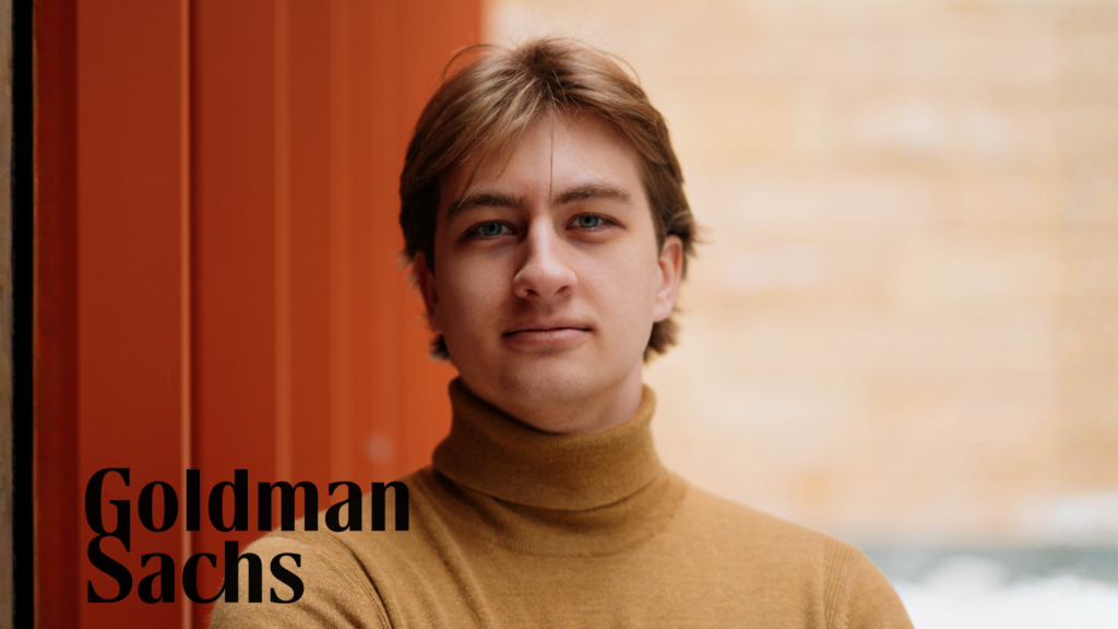 Dabney poses for a headshot in front of an orange background. He sports light brown hair, a tan turtleneck, and looks into the camera with a serious yet warm expression. The Goldman Sachs logo is displayed on the left hand side of the image.