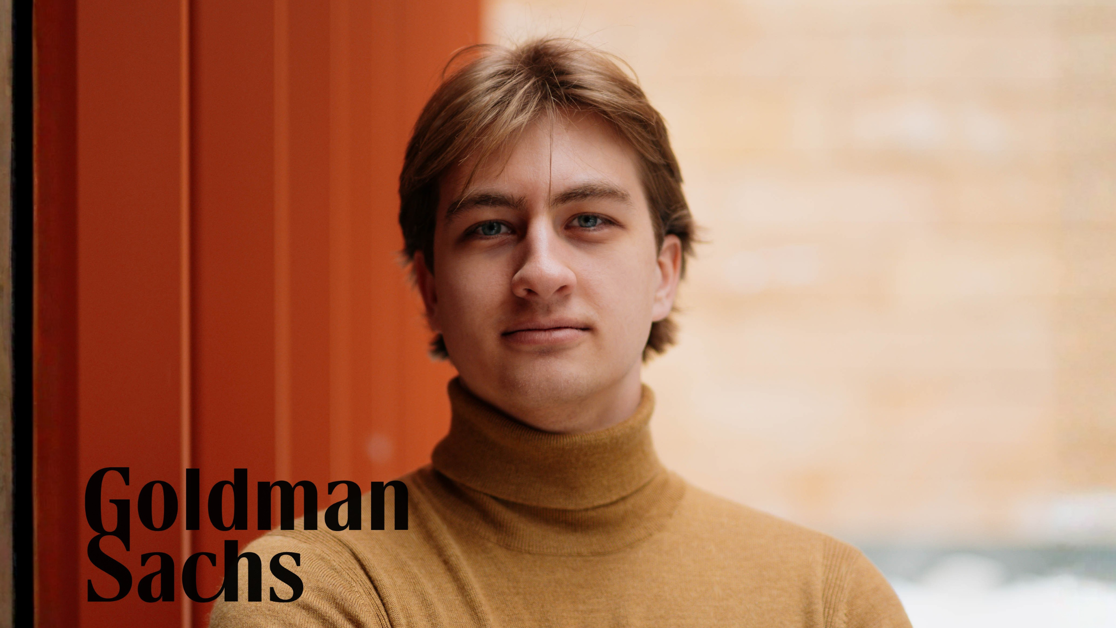 Dabney poses for a headshot in front of an orange background. He sports light brown hair, a tan turtleneck, and looks into the camera with a serious yet warm expression. The Goldman Sachs logo is displayed on the left hand side of the image.