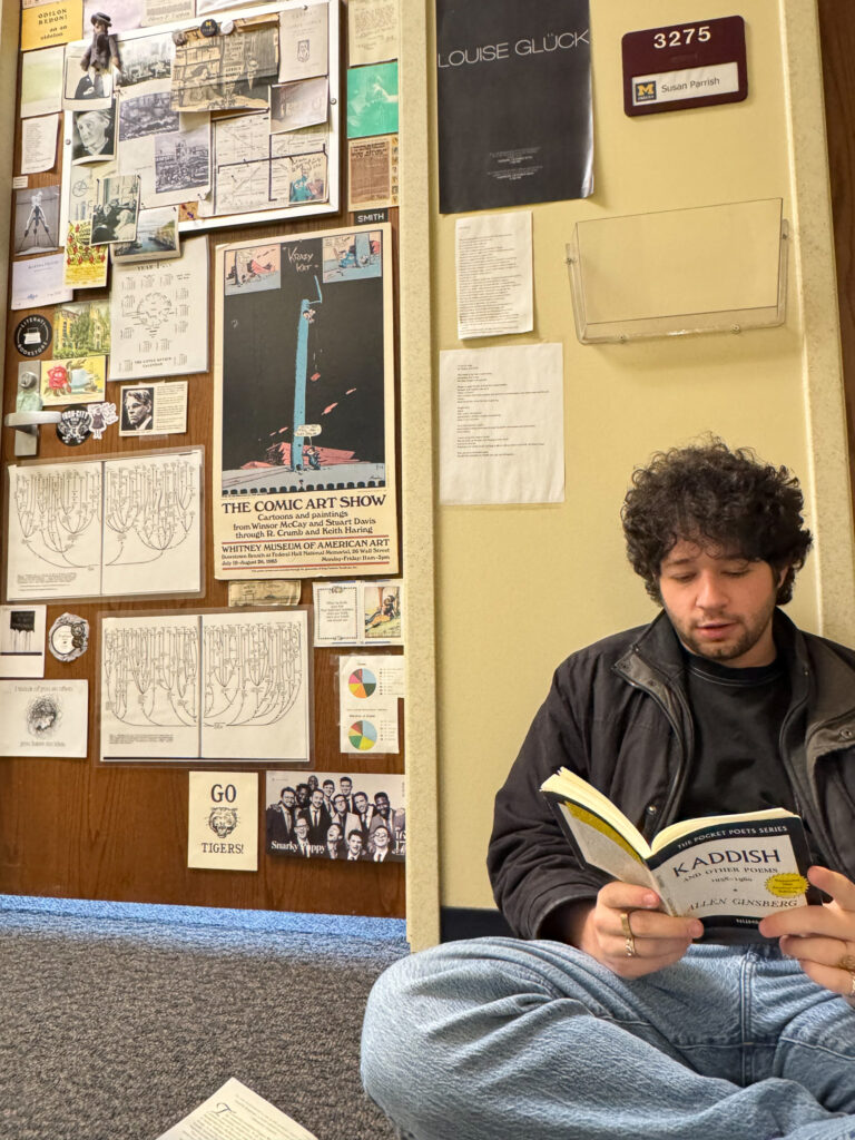 Max sits on the floor, reading a book of poetry outside what appears to be an office door at U-M.