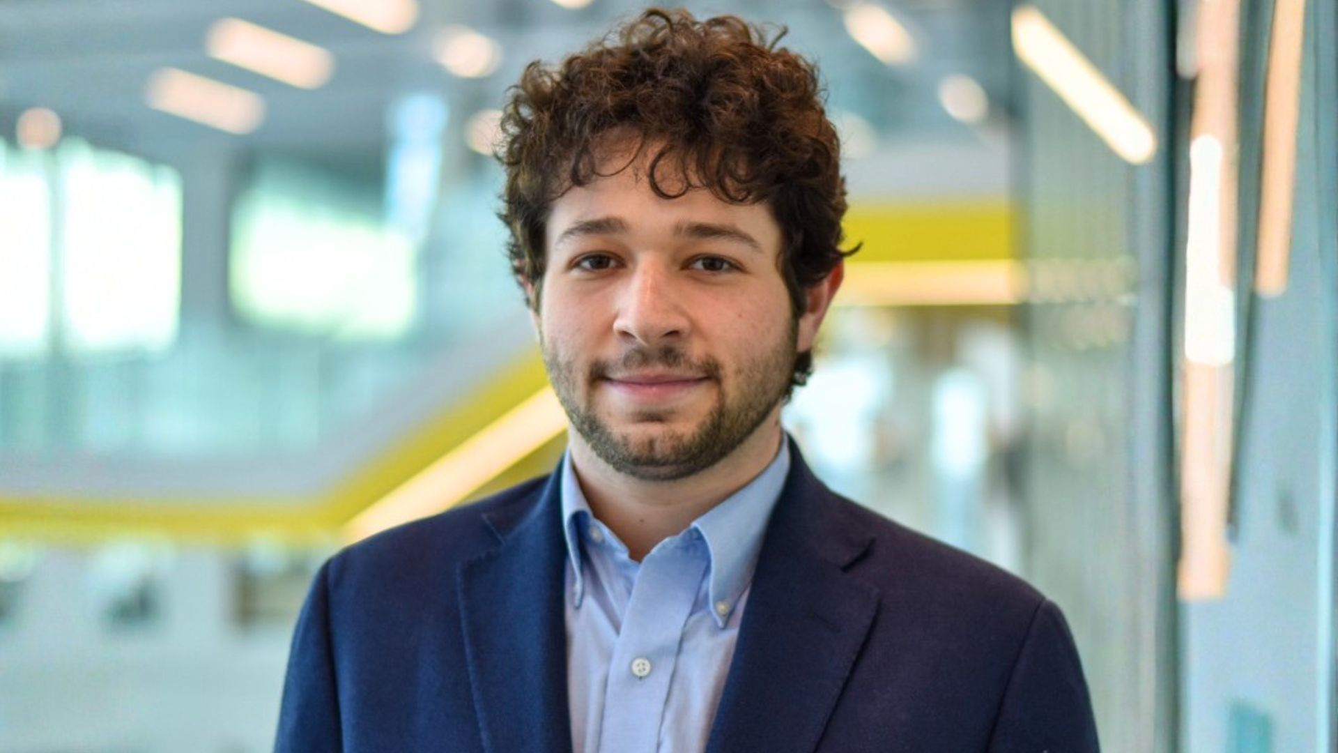 Max poses for a headshot in the Robotics building. He sports dark brown, curly hair, a beard, and a navy suit jacket with a light blue button-up shirt underneath.