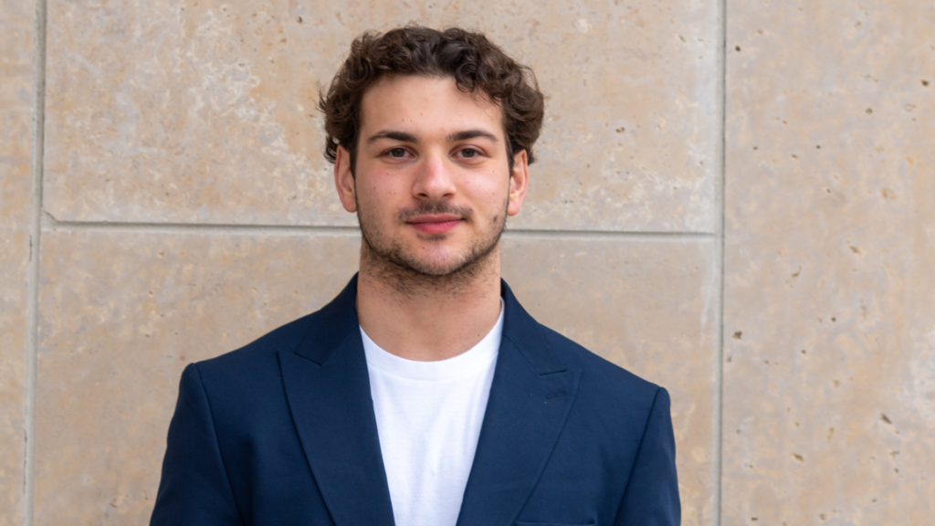 Michael poses for a headshot against a beige stone wall. He wears a navy jacket, white shirt, and has curly brown hair.