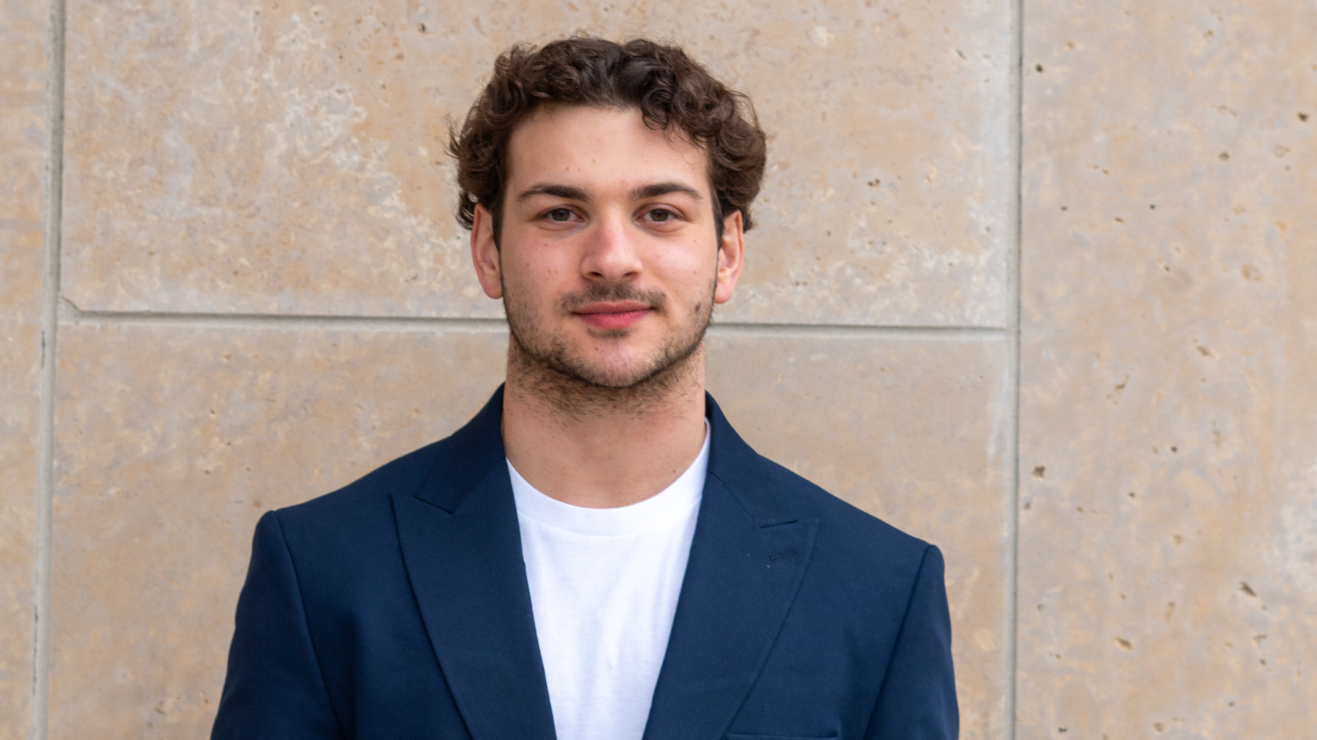 Michael poses for a headshot against a beige stone wall. He wears a navy jacket, white shirt, and has curly brown hair.