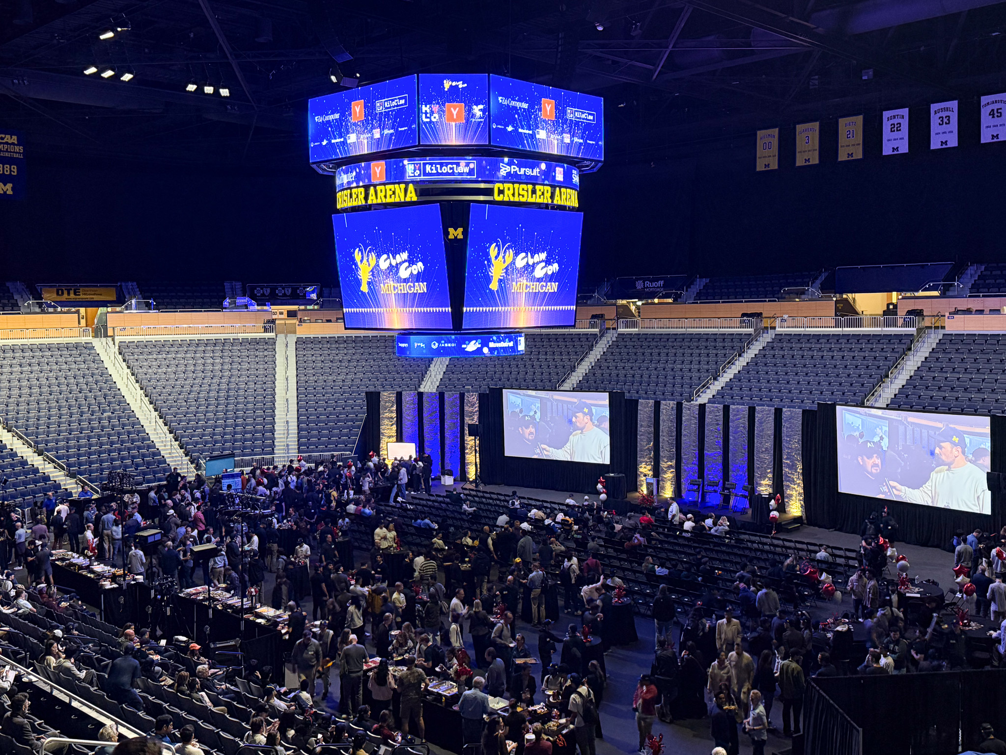 A scene from ClawCon Michigan at Crisler Arena in Ann Arbor Michigan. A ClawCon Michigan logo is displayed on the big screen at the arena and a large crowd is seated.
