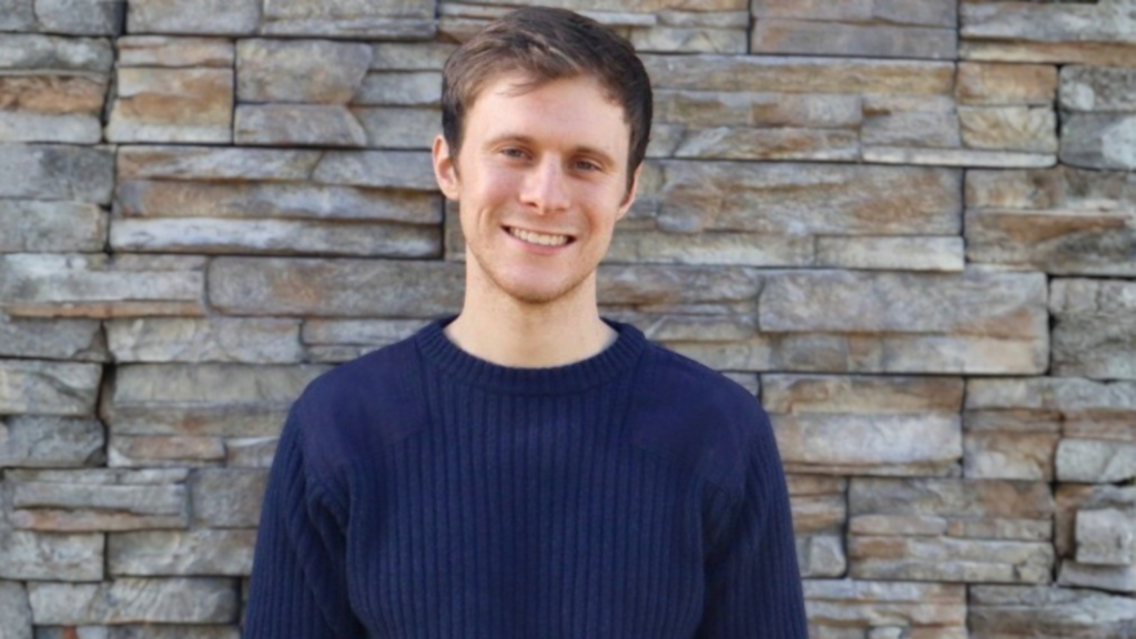 Clay Spaulding poses for a headshot against a grey stone background. He wears a navy sweater.