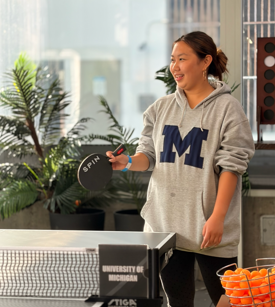 a woman in a Umich sweatshirt holding a ping-pong paddle
