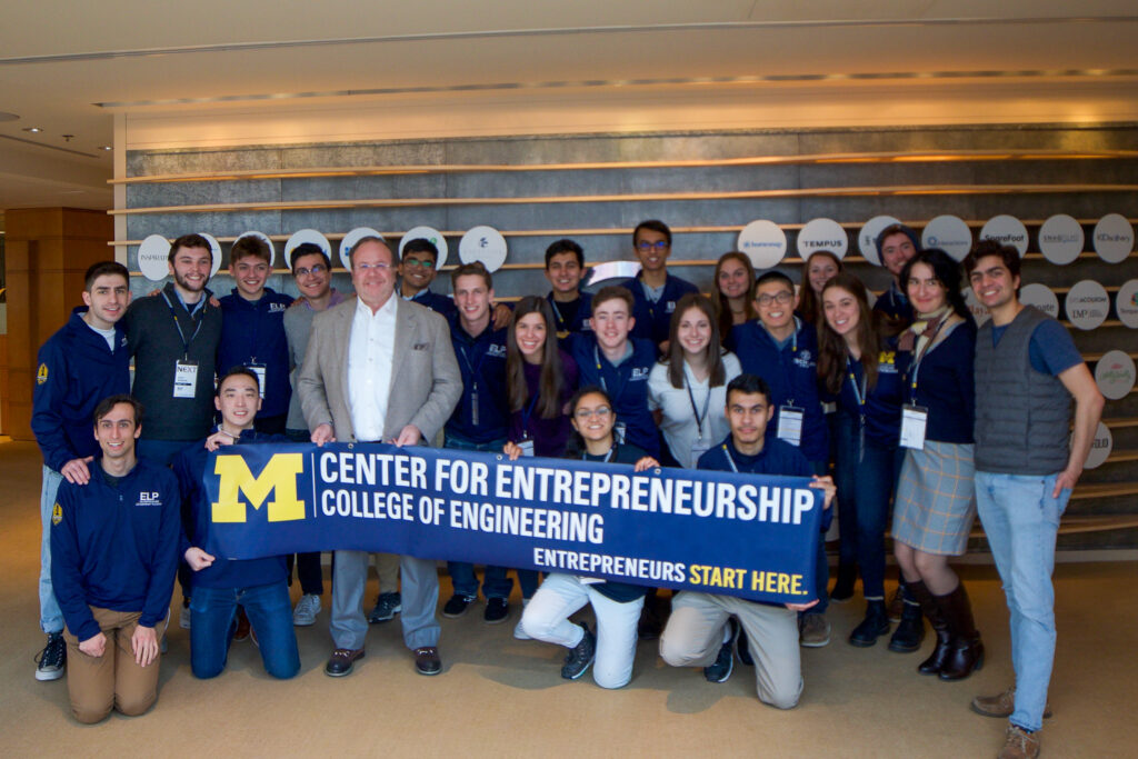Mira poses with fellow students on a CFE trek. The group holds a banner that reads "Michigan Center for Entrepreneurship. College of Engineering. Entrepreneurs start here.