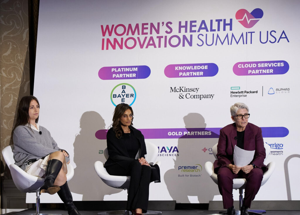 Mira speaks on a panel at the Women's Health Innovation Summit, along with two other women. They sit in chairs on a stage as they speak to the audience.