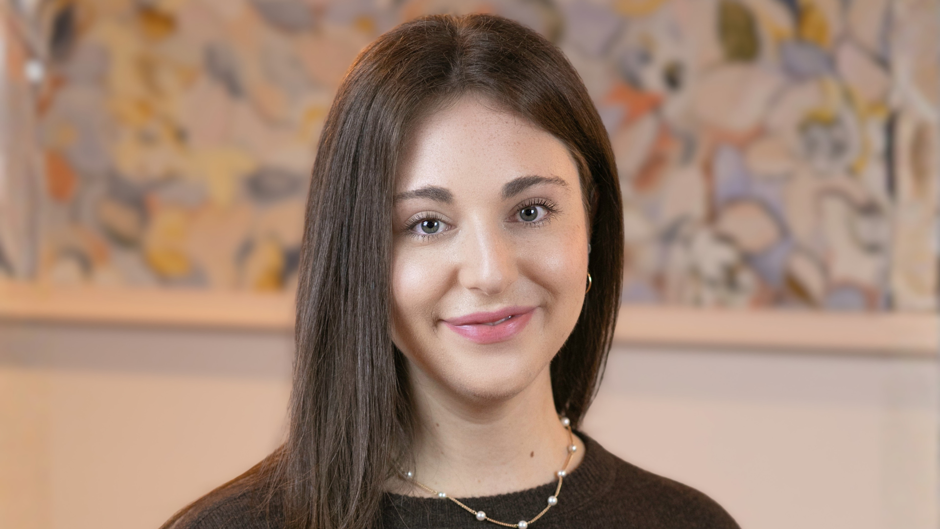 Mira Kaufman poses for a headshot. She has long, dark brown hair and gives the camera a slight smile. She stands against a wall with a floral wallpaper.