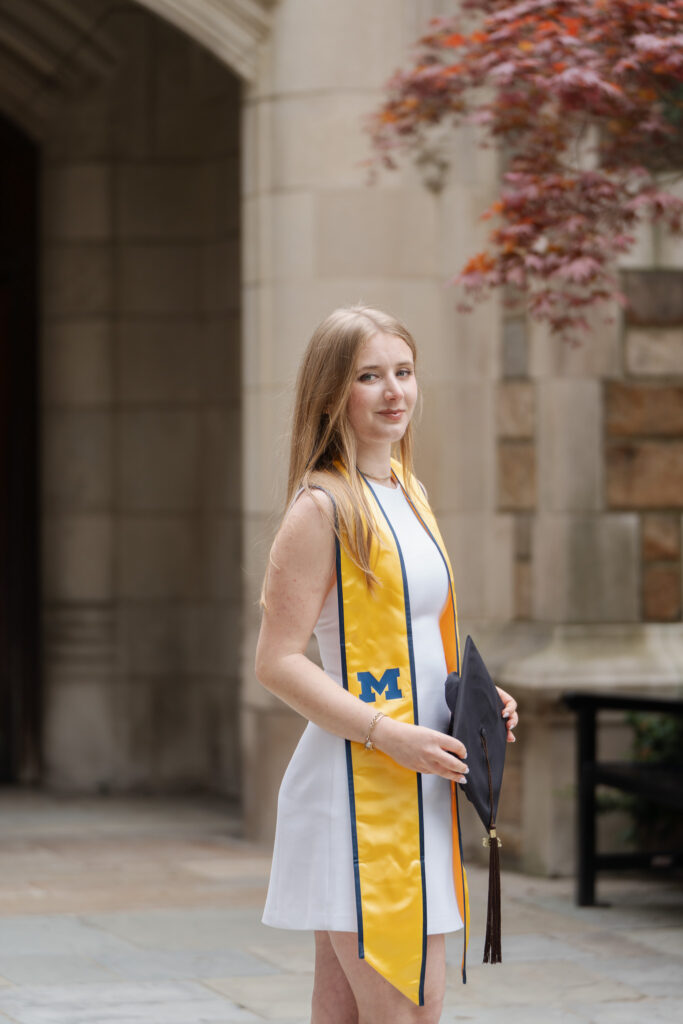 Julia Zislis poses in a white dress, holding a graduation cap in the U-M Law Quad.