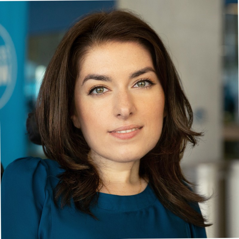 Mayaan Ziv poses for a headshot. She has medium length dark brown hair, brown eyes, and smiles at the camera wearing a dark blue shirt.