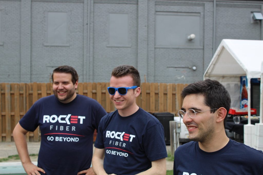 Ty is pictured standing with two colleagues, wearing a tshirt that reads "Rocket Fiber: Go beyond" and a pair of blue sunglasses. 