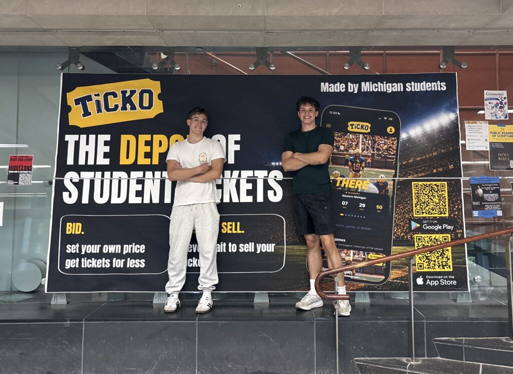 Isaac and co-founder Zach Devivo pose in front of a large banner advertising Ticko