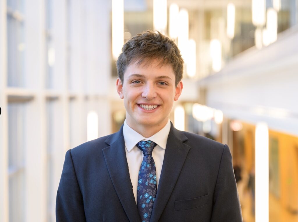 Isaac Gardner poses for a headshot. He wears a black suit and a blue tie.